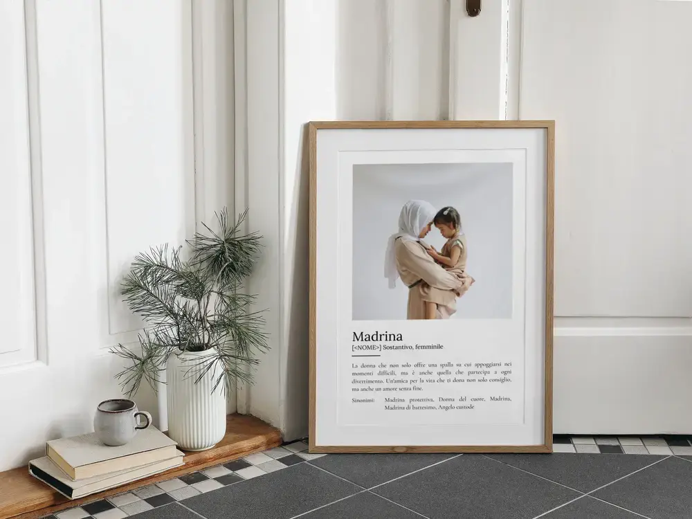 Poster per la madrina con foto e definizione, in cornice di legno appoggiata a terra accanto a libri e vaso