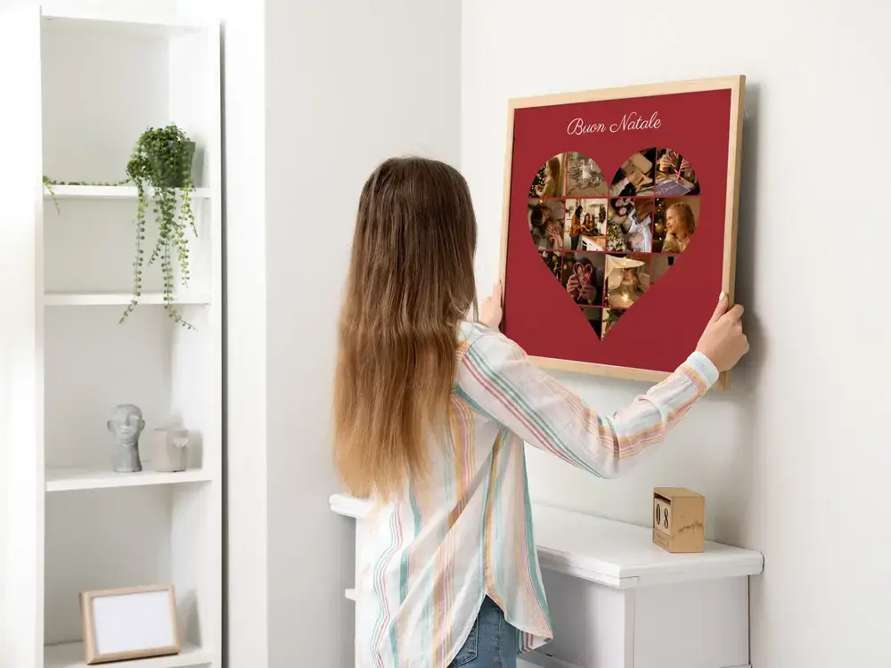 Collage di foto a cuore su fondo rosso con auguri di Natale, in cornice di legno chiaro su una parete bianca